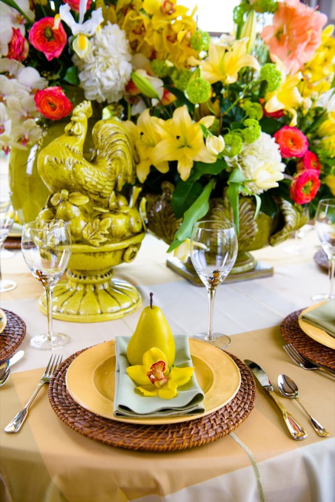Alamo Plants & Petals shows a yellow peach and flower atop a green napkin, atop a golden charger plate, atop a wooden wicker plate, atop a tabletop surrounded by yellow and red flowers with a golden rooster sculpture.
