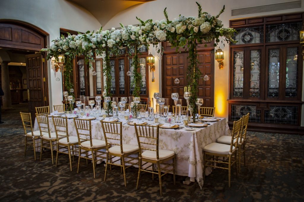 At The Dominion Country Club, in the center of the ballroomA long table for the wedding party is so elegant - there are glass goblets and gold chiavari chairs, with a lace white tablecloth all topped by a cover of flowers and florals.