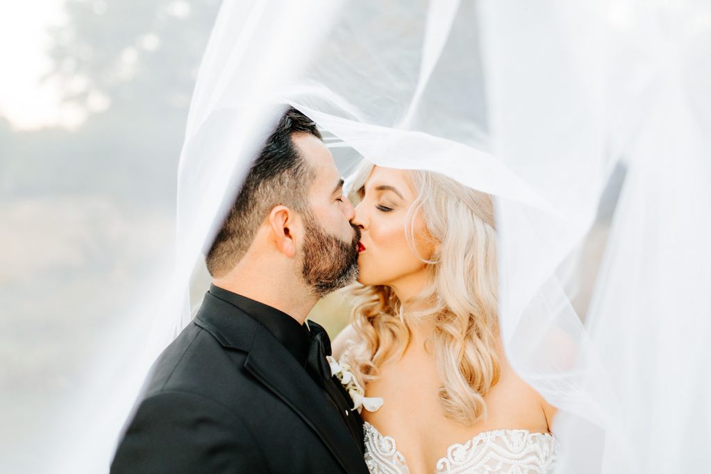newly wed couple kissing under the brides veil perspective shot